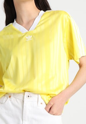Person, die ein gelbes Adidas-Shirt mit weißem Kragen und Streifen trägt, in weiße Hose mit hoher Taille gesteckt, eine Hand in der Tasche.