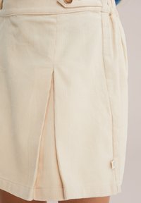 Cremfarbener Cordrock mit vorderem Faltenwurf, zwei seitlichen Taschen und einem kleinen Markenetikett am Saum. Mit einem Knopfverschluss gestaltet.