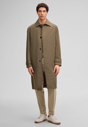 Mann trägt einen olivgrünen langen Knopfmantel, beige hochgekrempelte Hose und weiße Turnschuhe, steht vor einem einfarbigen Hintergrund.