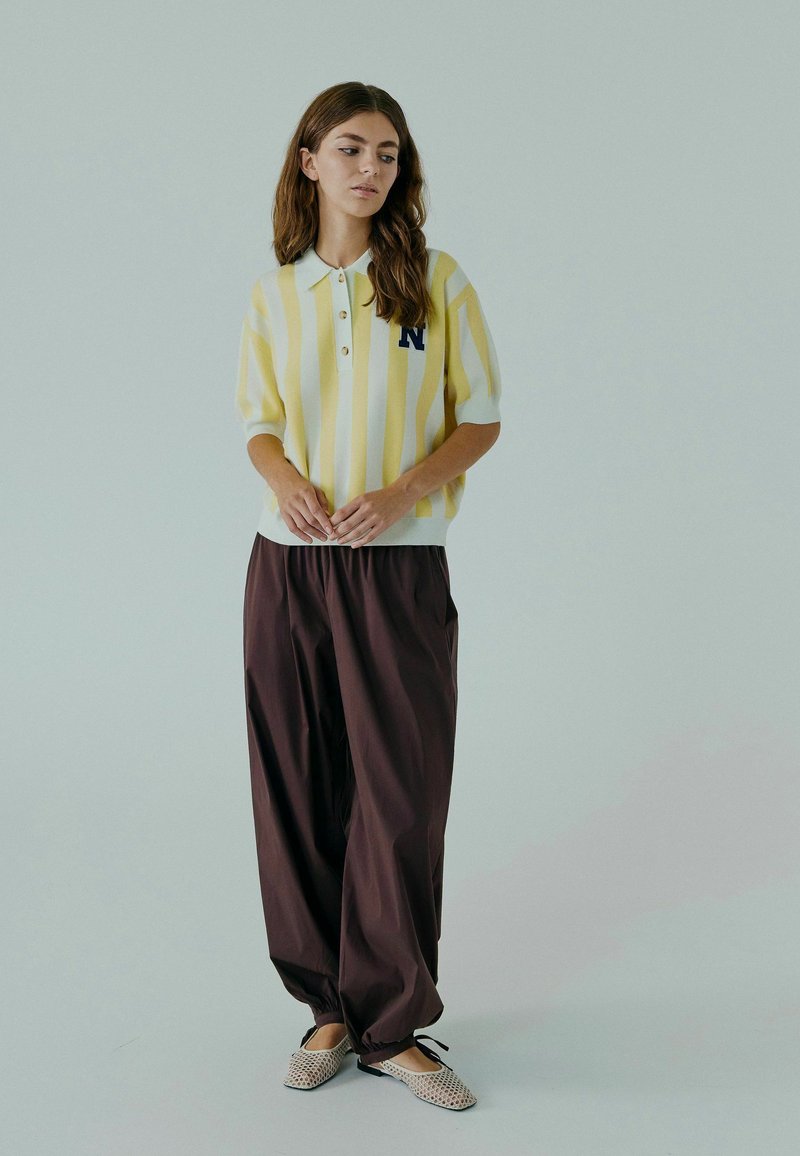 Polo tricoté avec des rayures jaunes et blanches, doté d'une patte de boutonnage à trois boutons et d'une lettre marine ; associé à un pantalon marron ample et des chaussures à motifs.