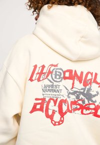 Felpa color crema con grandi stampe grafiche rosse e grigie sul retro, inclusi del testo e un design di una testa. Tessuto morbido e vestibilità comoda.