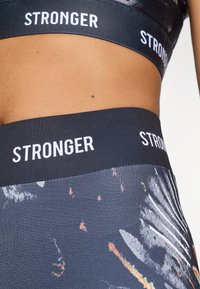 Black athletic crop top with white "STRONGER" branding and high-waisted leggings featuring a textured, abstract, multicolored pattern.