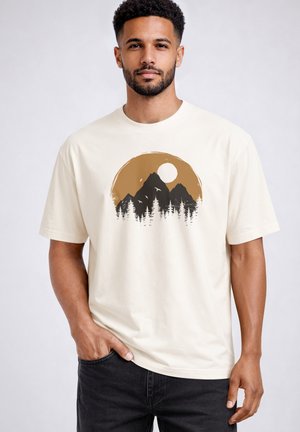 Man wearing cream T-shirt with mountain, pine trees, and sun graphic, standing with one hand in pocket against plain background.