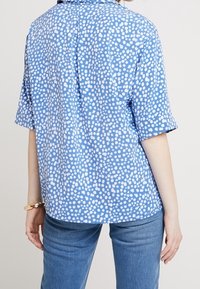 Blå kortärmad skjorta med vitt prickmönster, krage och avspänd passform. Materialet verkar slätt med en subtil lyster. Bakvy visas.