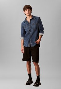 Junger Mann, der ein blaues Jeanshemd mit hochgekrempelten Ärmeln, schwarze knielange Shorts, schwarze Socken und schwarze Lederschuhe trägt.