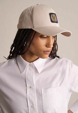 Femme aux cheveux tressés portant une casquette beige avec un patch foncé et une chemise blanche boutonnée avec une poche poitrine, regardant vers le bas sur le côté.