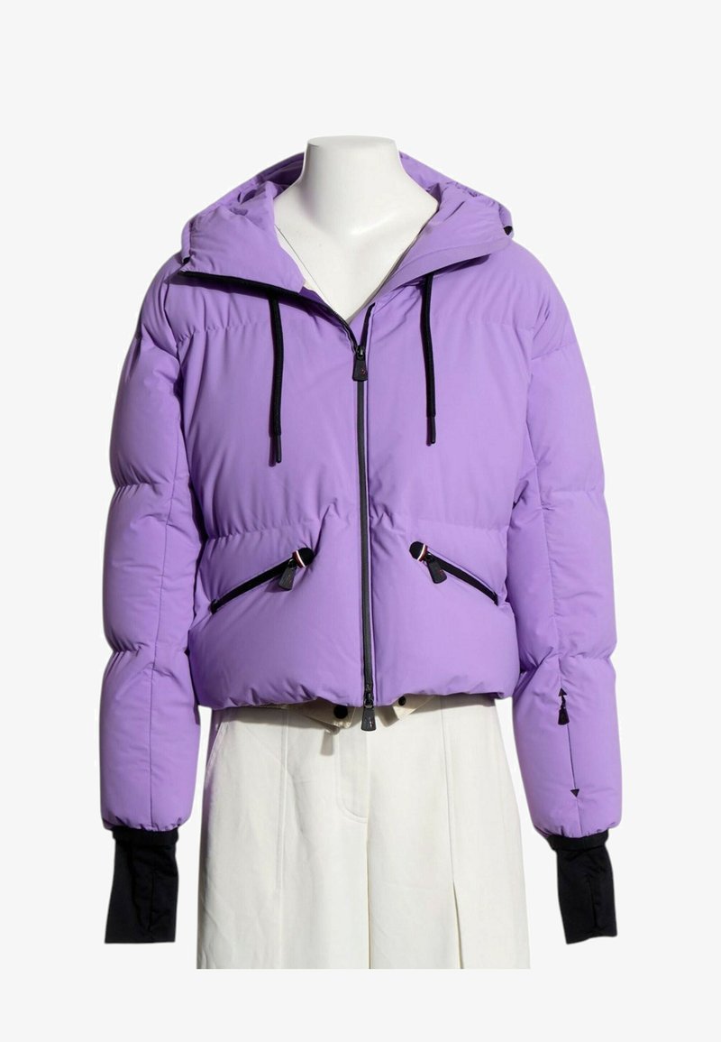 Doudoune violette avec capuche, cordons noirs, fermeture éclair à l'avant, deux poches zippées et poignets noirs allongés, portée sur un pantalon blanc.