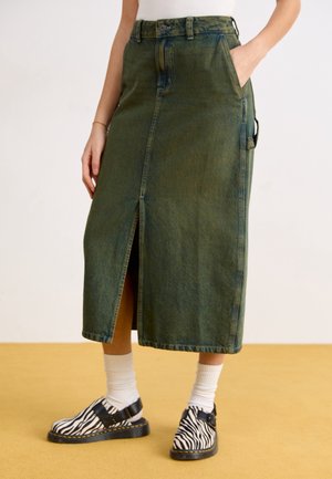 Persona con una falda midi de mezclilla verde con abertura frontal, calcetines blancos y zapatos con estampado de cebra en blanco y negro con correas de hebilla.