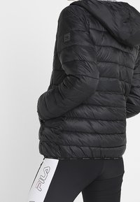 Svart puffjacka med huva, har horisontella quiltade sektioner, slät yta och en logotyp på vänstra axeln.