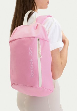 Ragazza che indossa uno zaino Reebok rosa chiaro con cerniera e cinghie bianche, vestita con una maglietta bianca e pantaloni beige, vista di profilo e da dietro.