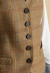 Hnedý károvaný vest s textúrovaného materiálu, obsahujúci päť tmavých gombíkov a dve bočné vrecká. Horizontálne a vertikálne pruhy v modrej a červenej farbe.