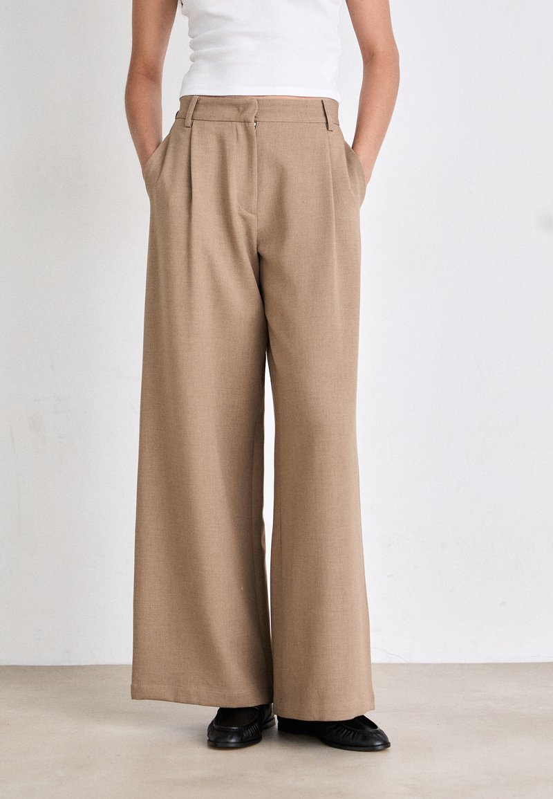Brede pantalon van lichtbruin stof met voorplooien, zijzakken en een getailleerde pasvorm. Draagt een fitting wit top en zwarte schoenen.