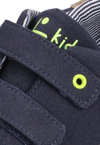 Zapato de lona azul con dos correas de velcro, detalle de ojales verdes y un forro interno a rayas. La marca está impresa en verde en la parte superior.