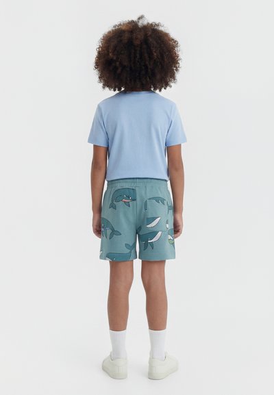 Göndör hajú gyermek áll háttal, világoskék pólót és türkiz rövidnadrágot visel, amelyen rajzfilm bálnák vannak, fehér zoknit és fehér cipőt.