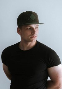 Tamna zelena snapback kapa s teksturiranim materijalom i detaljem loga u obliku dijamanta, u kombinaciji s prilagođenim crnim majicom na svijetloj pozadini.