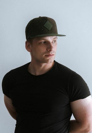Tamna zelena snapback kapa s teksturiranim materijalom i detaljem loga u obliku dijamanta, u kombinaciji s prilagođenim crnim majicom na svijetloj pozadini.
