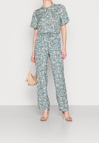 Completo floreale a due pezzi in tessuto blu-verde, caratterizzato da maniche corte, vita elasticizzata e pantaloni a gamba larga. Decorato con piccoli fiori bianchi.