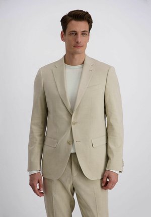 Homme portant un costume beige avec une veste et un pantalon, sur une chemise blanche, debout devant un fond gris clair uni.
