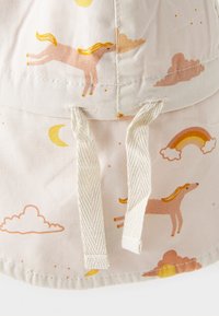 Gorra tipo bucket hecha de tela ligera que presenta un patrón de unicornios rosas, nubes y arcoíris sobre un fondo beige. Dos cordones en la parte delantera.