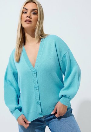 Kvinde iført en løs, lyseblå knappet cardigan og blå jeans, der står mod en ensfarvet hvid baggrund.