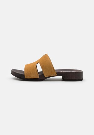 Sandal con unupper de ante amarillo mostaza y dos tiras recortadas, descansando sobre una suela de madera marrón oscuro con un tacón bajo.