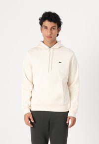 Sweat à capuche crème avec une poche kangourou et un cordon de serrage, arborant un petit logo vert sur la poitrine. Confectionné en tissu doux.