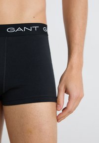 Männlicher Torso und oberer Oberschenkel in schwarzen GANT Boxershorts mit sichtbarem Taillenband-Logo, Hand ruht neben dem Oberschenkel auf einfarbigem Hintergrund.