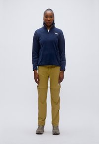 Μπλε ναυτικό φλις μπλουζάκι με φερμουάρ μέχρι το στήθος, συνδυασμένο με χακί παντελόνι καρότσα. Και τα δύο έχουν λογότυπα και άνετη εφαρμογή, φορεμένα με γκρι παπούτσια πεζοπορίας.