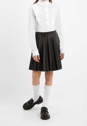 Kvinde iført en hvid flæset bluse, sort plisseret nederdel, hvide knæstrømper og sorte chunky loafers, stående på en hvid baggrund.