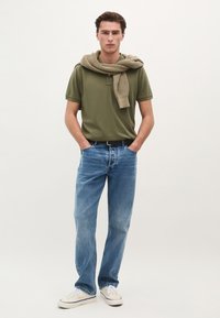 Grøn polo skjorte, beige striktrøje draperet over skuldrene, blå lige jeans og hvide sneakers. Simpelt design, afslappet pasform.