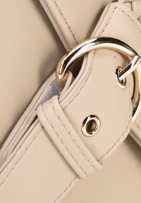 Beige konstläderväska med detaljer av en guldfärgad metallring och rem med stygn och en ögla. Fokus på den släta texturen.