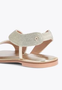 Sandal flat metallico dorato con una cinghia intrecciata testurizzata, una soletta liscia e una suola in gomma marrone chiaro. Accentata da una piccola gemma decorativa.
