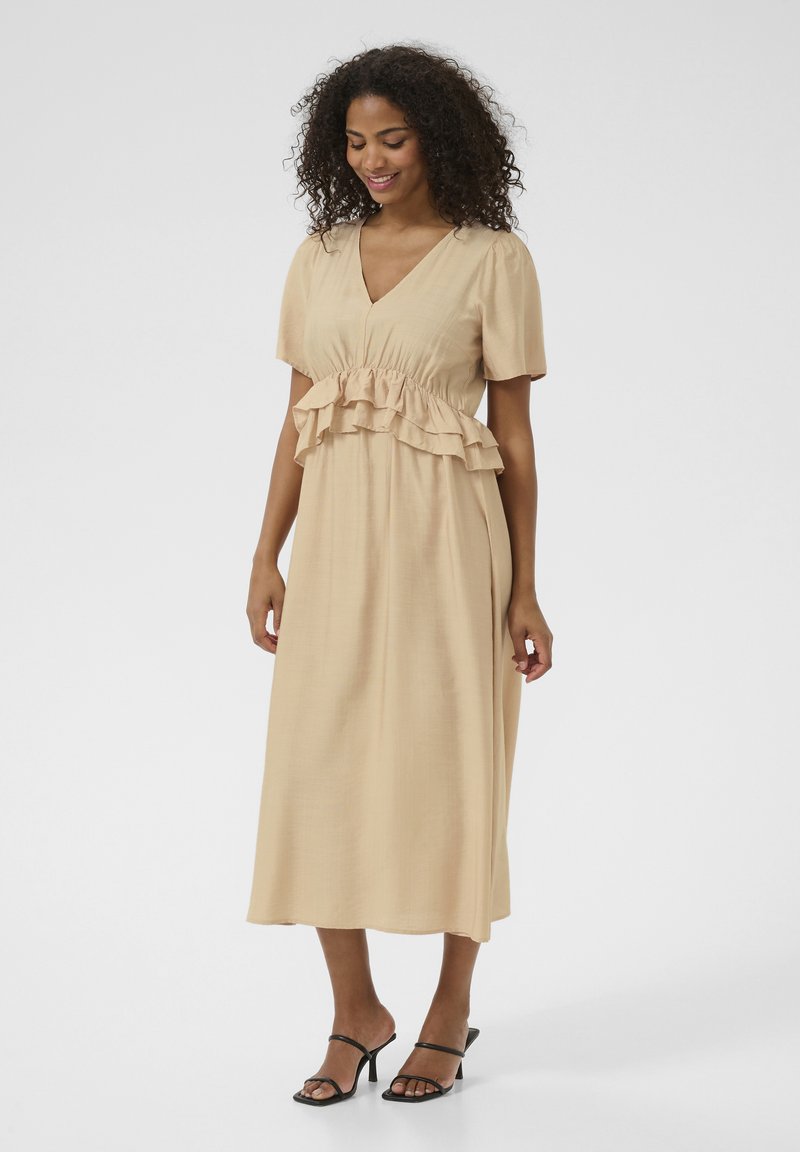 Beige midi-klänning med korta ärmar, volangdetaljer vid midjan och V-ringning. Slät textur, kombinerad med svarta sandaler med remmar.