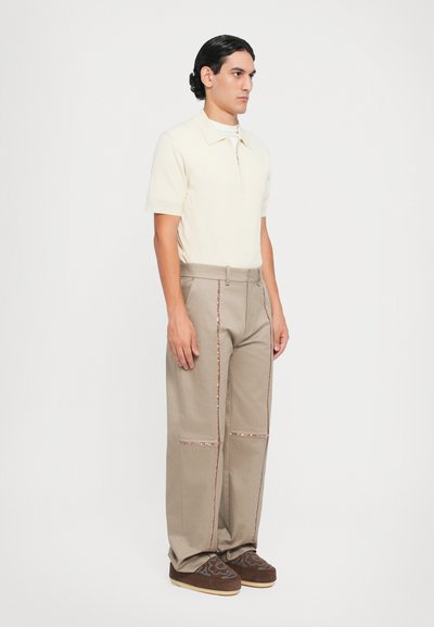Beige polo shirt met korte mouwen en een ritskraag; brede beige broek met koperen stiksels en zakken; bruine schoenen met patroon.