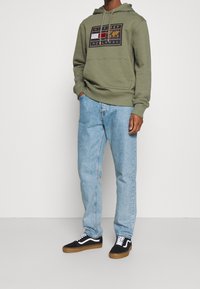 Felpa verde oliva con un design grafico, jeans in denim blu e sneakers nere con dettagli bianchi. Tessuto liscio e vestibilità rilassata.