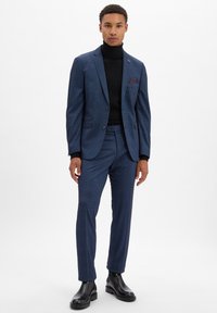 Marineblauer Anzug mit strukturierter Oberfläche, einreihige Jacke, schmale Hose, kombiniert mit einem schwarzen Rollkragenpullover. Leder-Ankle-Boots.