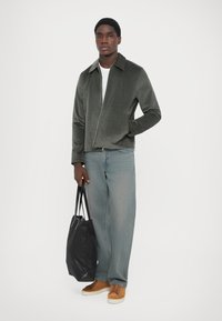Oliven grøn corduroy zip-jakke, hvid t-shirt, lyseblå jeans, brune ruskinds sneakers, sort tote bag. Simpelt design med krave og lommer.