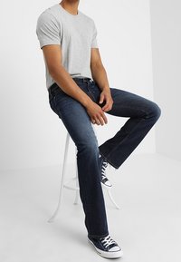 Camiseta de algodón gris, jeans de mezclilla azul oscuro, ajuste entallado. Lleva zapatillas altas en blanco y negro. Hombre sentado en un taburete blanco.