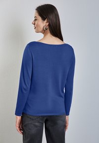 Blauer Langarmpullover mit weitem Rundhalsausschnitt und gerippten Bündchen. Weicher Stoff, figurbetonter Schnitt, kombiniert mit dunkelgrauen Jeans.