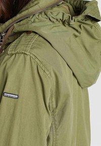 Veste à capuche vert olive avec texture froncée, matériau léger, fermeture éclair visible et écusson logo sur l'épaule gauche.