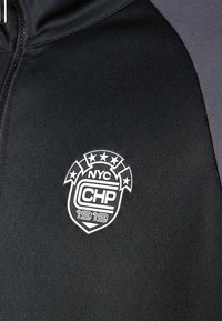 Juoda striukė su baltu NYC CHP 1919 emblematu, kuriame viršuje yra penkios žvaigždės ir stilizuoto skydelio dizainas ant krūtinės.