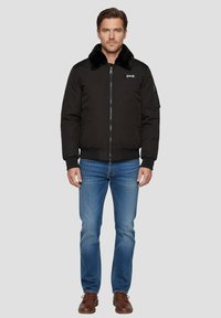 Giacca bomber nera con colletto in pelliccia sintetica, zip frontale, polsini e orlo a coste. Abbinata a jeans blu e scarpe marroni, il modello è in piedi su uno sfondo grigio.