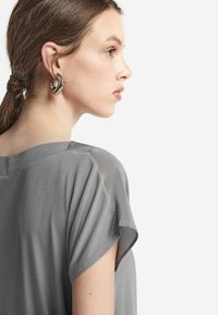 Grå blus med kort ärm i mjukt, lätt genomskinligt material. Modellen bär en distinkt silverörhänge som visar en samtida design och form.