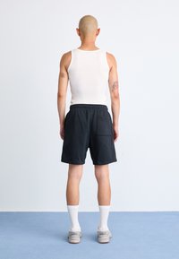 Sorte atletiske shorts med elastisk talje, iført en hvid tanktop og grå sneakers, hvide ankelsokker, set fra ryggen.