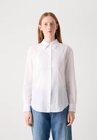 White long-sleeve shirt with a pointed collar and button closure, made of lightweight fabric, paired with blue high-waisted denim.
