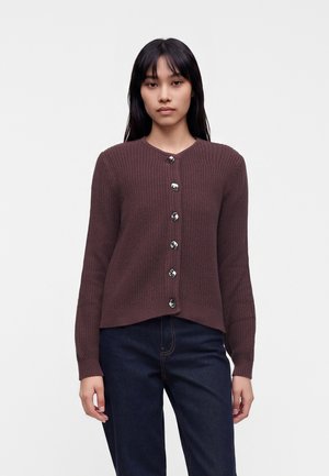 Femme aux longs cheveux foncés portant un cardigan marron en laine boutonné et un jean bleu foncé, debout devant un fond clair uni.