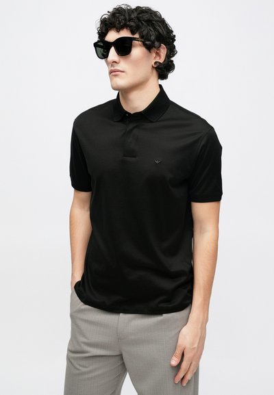 Melnas polo krekls no gludas auduma, ar klasisku apkakli un īsām piedurknēm. Valkāts kopā ar pelēkiem rūtainiem biksēm un tumšām saulesbrillēm.