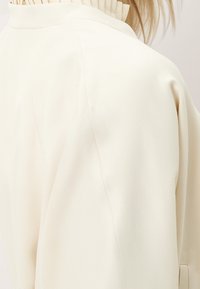 Giacca bianca strutturata con colletto a coste, caratterizzata da un tessuto leggermente testurizzato e cuciture che esaltano il suo design sartoriale.