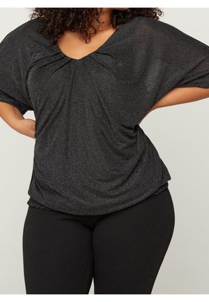 Person med krøllet hår iført glitrende sort V-hals top og sorte leggings med hænderne hvilende på hoften mod en ensfarvet baggrund.