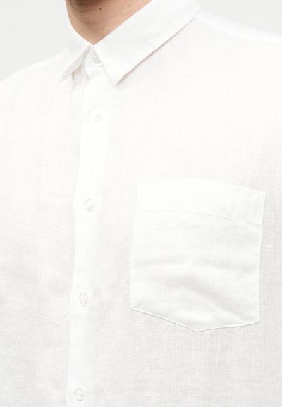 Κοντινό πλάνο ενός ατόμου που φοράει λευκό πουκάμισο με κουμπιά, με τσέπη στο στήθος και μυτερό γιακά, πάνω σε απλό φόντο.